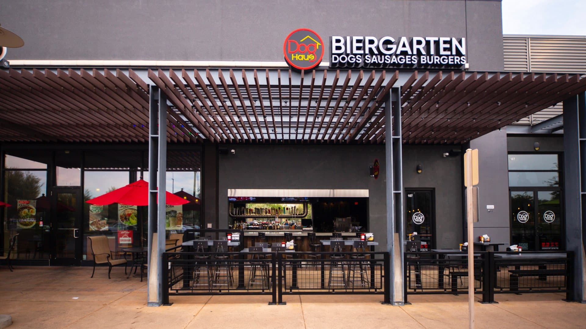 An outdoor patio of a restaurant with a bar visible through a large opening, featuring tables, chairs, and a green umbrella.